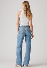 Light blue denim jeans with a wide-leg fit, featuring two back pockets and a leather logo patch at the waistband. White tank top and sneakers worn.