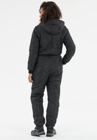 Zwarte gewatteerde jumpsuit met een capuchon, elastische taille en ribbelmanchetten. De stof heeft een gladde textuur en vertoont diagonale stikpatronen.