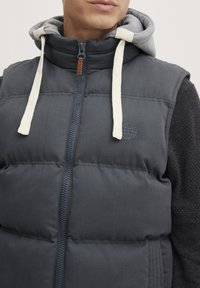 Mørk grå dunvest med høy krage, lysegrå hette, to-farget snorer og en subtil logo. Teksturerte sidesegmenter tilfører detaljer.