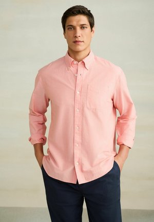 Mladý muž stojaci s rukami v vreckách, oblečený v svetloružovej košeli na gombíky a tmavomodrých nohaviciach, na neutrálnom pozadí.