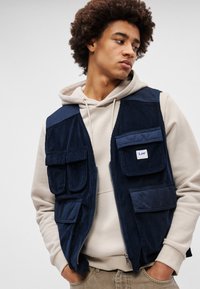 Gilet in corduroy blu navy con diverse tasche frontali, chiusura con zip e logo a patch. Indossato sopra un cappuccio di colore beige chiaro.