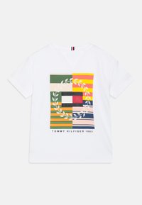 Camiseta de algodón blanca con un gráfico colorido que presenta rayas y un diseño de hoja circular, etiquetada como "TOMMY HILFIGER 1985" en la parte inferior.