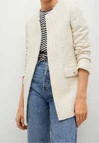 Witte tweed jasje met textuurstof, open voorkant en subtiele zakaccenten. Gedragen over een zwart-wit gestreept shirt en blauwe jeans.