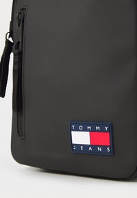 Black nylon backpack featuring a front zipper pocket, textured surface, and a branded patch in blue, red, and white with "TOMMY JEANS."