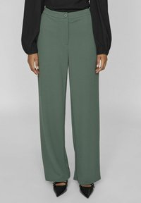 Pantalones de pierna ancha en color verde salvia, hechos de una tela suave; cuentan con un cierre de botón y pliegues en la parte frontal, combinados con zapatos negros de punta.