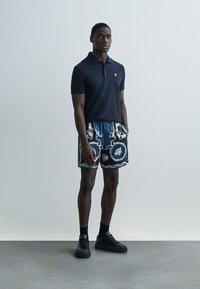 Tmavomodrá polokošeľa, vzorované modro-čierne šortky s ozdobnými motívmi a čierne tenisky. Model stojí na svetlom pozadí.