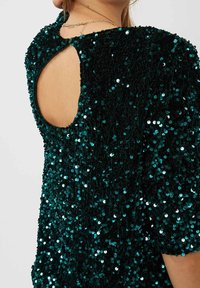 Top verde petrolio con paillettes e maniche corte, caratterizzato da un tessuto testurizzato e un'apertura ovale sul retro, che rivela la pelle nuda sottostante.