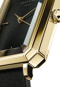 Reloj rectangular en tono dorado con una esfera negra texturizada, que presenta manecillas doradas de hora y minuto, y una correa de cuero. Destaca por su detalle de corona estriada.
