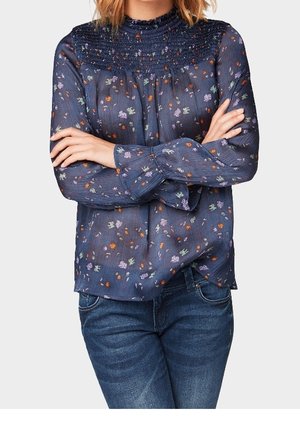 Femme les bras croisés portant un chemisier bleu marine à fleurs à manches longues avec un col smocké et un jean bleu.