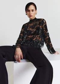 Top nero trasparente a maniche lunghe con design in pizzo floreale, colletto alto e ricami elaborati. Abbinato a pantaloni in satin neri a vita alta.