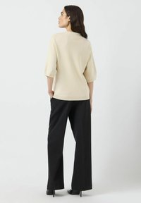 Beige, löst sitzendes Strickoberteil mit kurzen Ärmeln, kombiniert mit schwarzen weiten Hosen und spitzen schwarzen Absatzschuhen, von hinten gesehen.