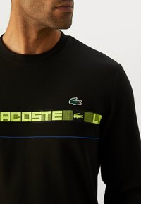 Svart hettegenser med rund hals, fluorescerende grønn grafikktrykk over brystet, og en liten grønn krokodillelogo på venstre side.