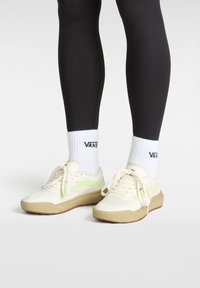 Cream and green sneakers with a textured surface, featuring a chunky light brown sole and laces, paired with white socks displaying a logo.