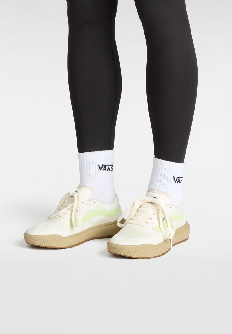 Cream and green sneakers with a textured surface, featuring a chunky light brown sole and laces, paired with white socks displaying a logo.