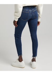 Jeans de denim azul ajustados con un dobladillo deshilachado, que cuentan con dos bolsillos traseros y una cintura alta, combinados con zapatillas blancas.