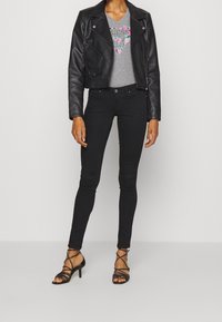 Kvinde iført en sort cropped læderjakke, grå grafisk t-shirt, sorte stramme bukser og sorte højhælede sandalstøvler, stående foran en ensfarvet baggrund.