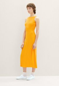 Ein ärmelloses, knöchellanges Kleid aus leuchtend orangefarbenem Stoff, mit einem taillierten Oberteil und gerader Silhouette. Kombiniert mit weißen Turnschuhen.