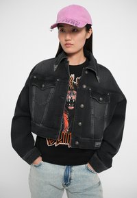 Črna skrajšana jeans jakna s srebrnimi gumbi, z dvema žepoma na prsih, nosi se preko črne majice s grafiko, ki prikazuje oranžnega tigra. Svetle jeans hlače.