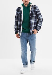 Camicia flanella a quadri in navy, blu e bianco con un morbido rivestimento in pile bianco. Abbinata a una felpa verde e jeans azzurri chiari. Sneakers bianche.