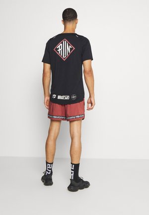 Hombre de pie de espaldas, vistiendo una camiseta negra con el gráfico "RUN", pantalones cortos rojos para correr con ribetes negros y blancos, calcetines negros y zapatillas deportivas negras.