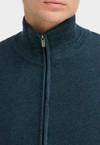 Knitted zip-up sweater in dark teal, featuring a high collar, textured fabric, and a sleek front zipper with a metallic pull.
