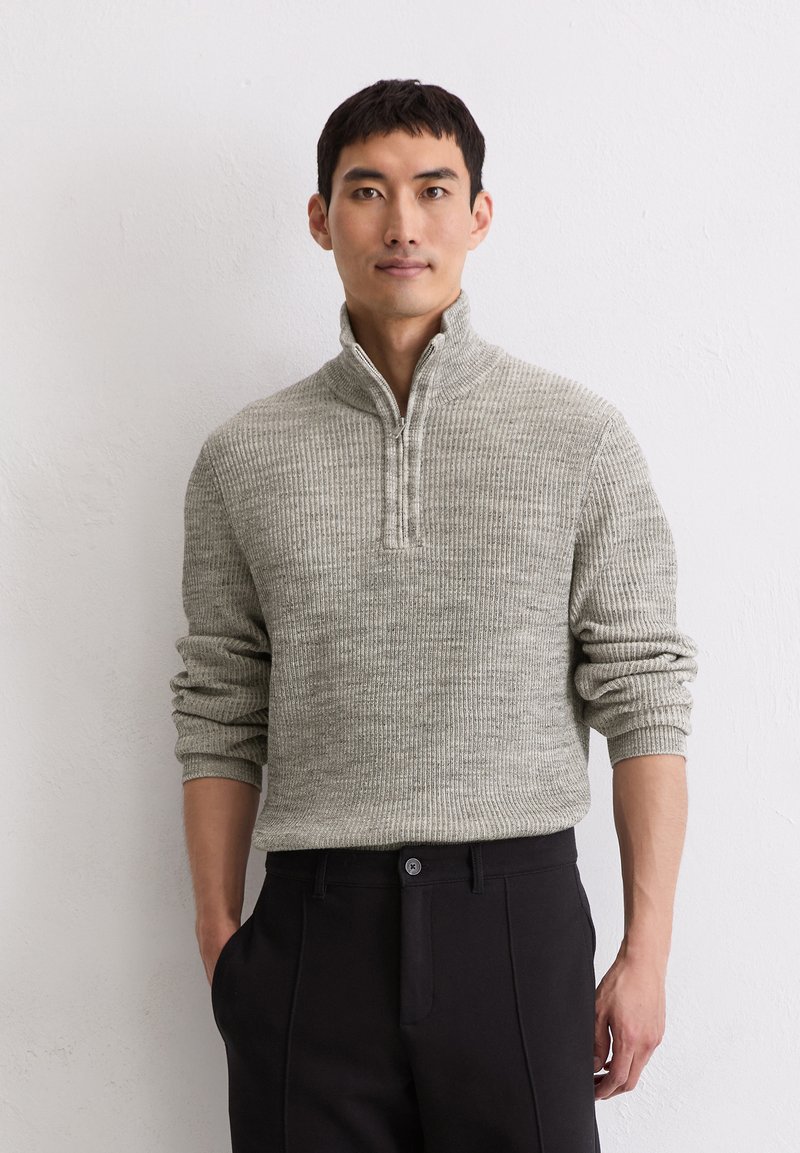 Hellgrauer, gerippter Pullover mit hohem Kragen und einem Viertelreißverschluss. Kombiniert mit dunklen Hosen, die eine strukturierte Passform und Textur aufweisen.