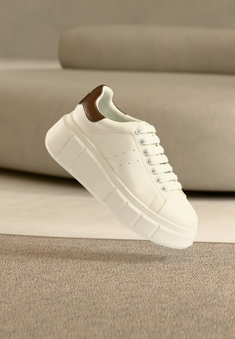 Λευκό δερμάτινο αθλητικό παπούτσι με καφέ δερμάτινη λεπτομέρεια, χοντρή σόλα, στρογγυλή μύτη και επίπεδα κορδόνια. Διαθέτει διακριτικές τρύπες στη πλευρά.