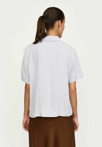 Krótki rękaw, jasno szara bluzka z kołnierzykiem, gładka tekstura, luźny krój i zaokrąglony dół. Noszona z brązową spódnicą.