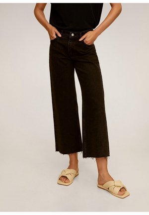 Osoba nosząca czarne, szerokie, krótkie jeansy z wysokim stanem, z surowymi brzegami oraz beżowe, plecione klapki na płaskiej podeszwie, ręce w kieszeniach.