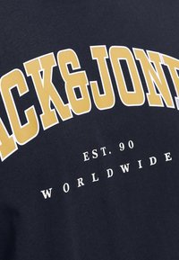 Navy cotton T-shirt featuring large white and yellow "JACK & JONES" lettering, with "EST. 90" and "WORLDWIDE" printed below in white.