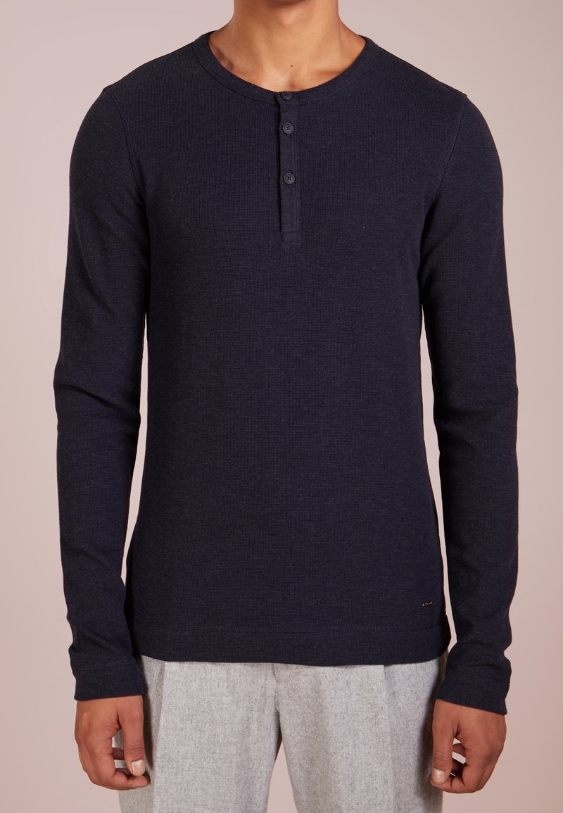 Man wearing a fitted dark navy long-sleeve Henley shirt with three buttons, paired with light gray pants, neutral background.