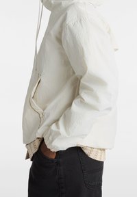 Vit, lättviktig jacka med texturerad yta, huva, ficka med dragkedja och elastiska ärmslut, lagd över en mönstrad skjorta och svarta jeans.