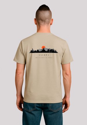 Man with shaved mohawk wearing beige t-shirt with mountain and city skyline graphic and orange sun on back, paired with blue jeans.