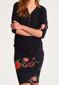 Black blouse with a zip neckline, paired with a fitted black skirt featuring multicolored floral patterns and birds. Smooth texture, vibrant colors.