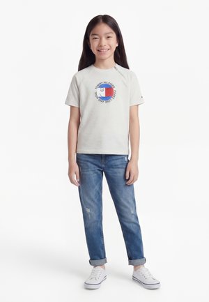 Κοπέλα με μακριά σκουρόχρωμα μαλλιά που χαμογελά, φοράει λευκό T-shirt με σχέδιο, μπλε τζιν με ρεβερ, και λευκά αθλητικά παπούτσια, στέκεται μπροστά σε λευκό φόντο.