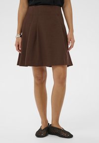 Femme portant une jupe plissée à carreaux bruns, un haut noir, un bracelet en argent et des chaussures plates tissées noires, se tenant devant un fond uni.