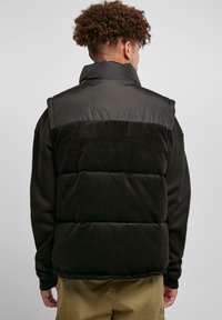 Vest puf cu catifea neagră și țesătură lucioasă, guler înalt, textură matlasată și o croială lejeră, purtat peste un tricou cu mâneci lungi de culoare închisă.