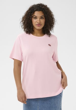 Kvinde med krøllet hår iført en lyserød t-shirt med et lille brunt blomsterlogo og et blåt denimsnørklæde mod en hvid baggrund.