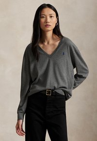 Kvinne med langt mørkt hår har på seg en løs grå V-hals genser og svarte bukser med svart belte, står foran en ensfarget beige bakgrunn.