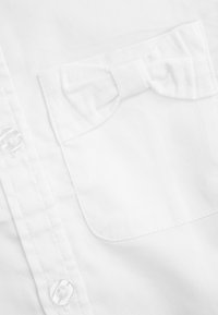 Biała koszula z guzikami, wykonana z fakturowanej tkaniny, z kieszonką na lewym piersi i akcentem w formie kokardy oraz przezroczystymi plastikowymi guzikami.