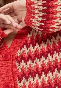 Cardigan en tricot rouge avec un motif en zigzag en crème et différentes nuances de rouge. Comprend de grands boutons rouges et des manches texturées.