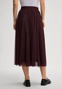 Woman wearing a dark burgundy midi tulle skirt with an elastic waistband paired with black leather loafers, shown from the back.
