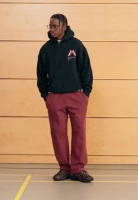 Fekete kapucnis pulóver vörös grafikával, bordó nadrág, és fekete cipők többféle textúramintával. Fa fal előtt.