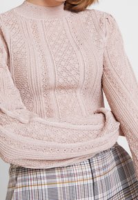 Koronkowy sweter z długim rękawem w jasnoróżowym kolorze z teksturowanymi wzorami i wysokim kołnierzem. Połączony z kratkowaną spódnicą z czarnymi, białymi i żółtymi liniami.