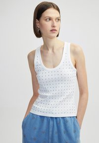 Weißes Tanktop mit einem runden Ausschnitt, das ein gepunktetes Muster aus kleinen schwarzen Verzierungen aufweist. Gerippte Textur; figurbetontes Design.