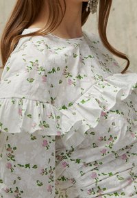 Blusa blanca de flores con estampado de flores rosas y hojas verdes, con mangas acampanadas y una textura fruncida. Tejido suave con un patrón delicado.