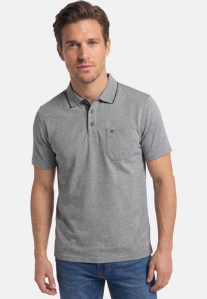 Mann mit kurzen braunen Haaren, der ein graues kurzärmliges Poloshirt mit Brusttasche und blaue Jeans trägt, steht vor einem einfarbigen Hintergrund.