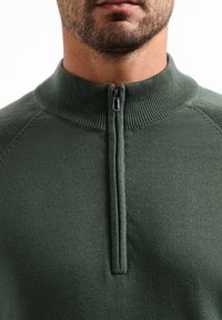 Groene ritspullover met een hoge kraag, ribbelachtige halslijn en een gladde textuur. De rits is centraal geplaatst aan de voorkant.