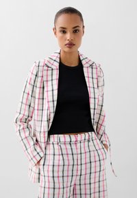 Tweedelige pak in lichte stof met een roze en zwarte ruitpatroon. De blazer heeft een geknipte kraag, terwijl de broek op maat is gemaakt met zakken.