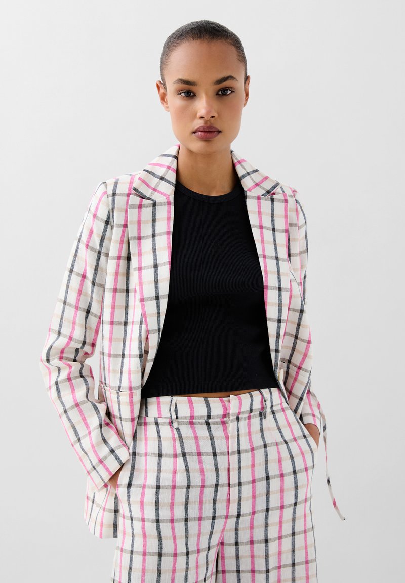 Tweedelige pak in lichte stof met een roze en zwarte ruitpatroon. De blazer heeft een geknipte kraag, terwijl de broek op maat is gemaakt met zakken.